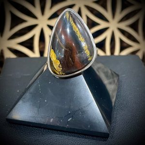 Bague argent et pierre naturelle en œil de fer de forme triangulaire. Photo de face, bague posée sur une pyramide noire sur fond noir et fleur de vie en bois ajouré