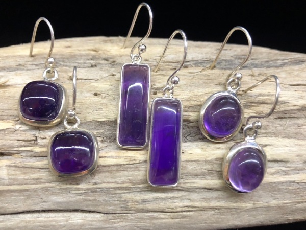 Trois paires de boucles d'oreilles en améthyste présentées sur un morceau de bois flotté sur fond noir