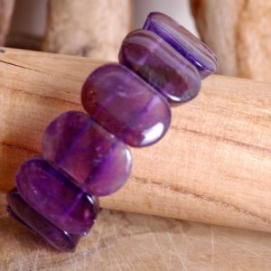 Bracelet améthyste perles plaquettes de forme rectangulaire. Il présenté de travers sur un rondin de bois sur fond bois