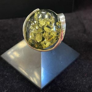 Bague ronde en argent avec une pierre minérale naturelle d’ambre vert de grosse taille. Montage atypique. Photo prise de face sur fond noir avec la bague posée sur une pyramide de Shungite noire