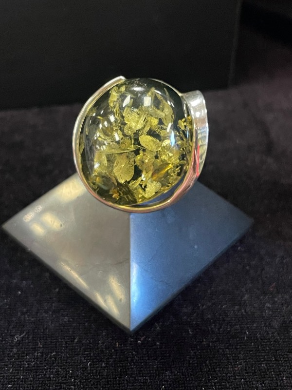 Bague ronde en argent avec une pierre minérale naturelle d’ambre vert de grosse taille. Montage atypique. Photo prise de face sur fond noir avec la bague posée sur une pyramide de Shungite noire