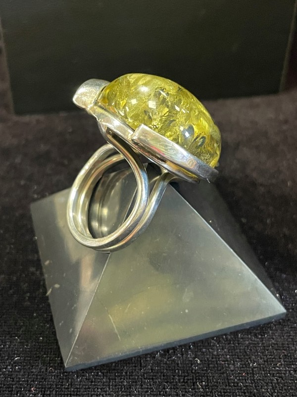 Bague ronde en argent avec une pierre minérale naturelle d’ambre vert de grosse taille. Montage atypique. Photo prise de face sur fond noir avec la bague posée sur une pyramide de Shungite noire