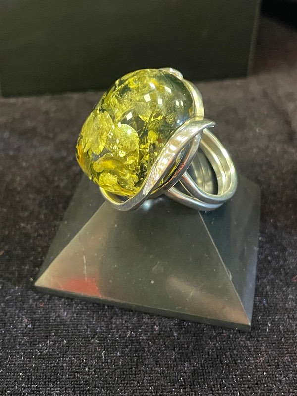 Bague ronde en argent avec une pierre minérale naturelle d’ambre vert de grosse taille. Montage atypique. Photo prise de profil sur fond noir avec la bague posée sur une pyramide de Shungite noire