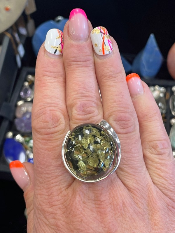 Bague ronde en argent avec une pierre minérale naturelle d’ambre vert de grosse taille. Montage atypique. Photo prise de face avec la bague portée sur une main, gros plan sur la bague sur fond noir