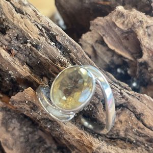 Bague argent et citrine ovale Faceté. Montage atypique avec l’anneau qui part d’un côté de l’oval pour rejoindre l’autre extrémité de l’oval. Photo de dessus, bague posée sur un morceau de bois flotté