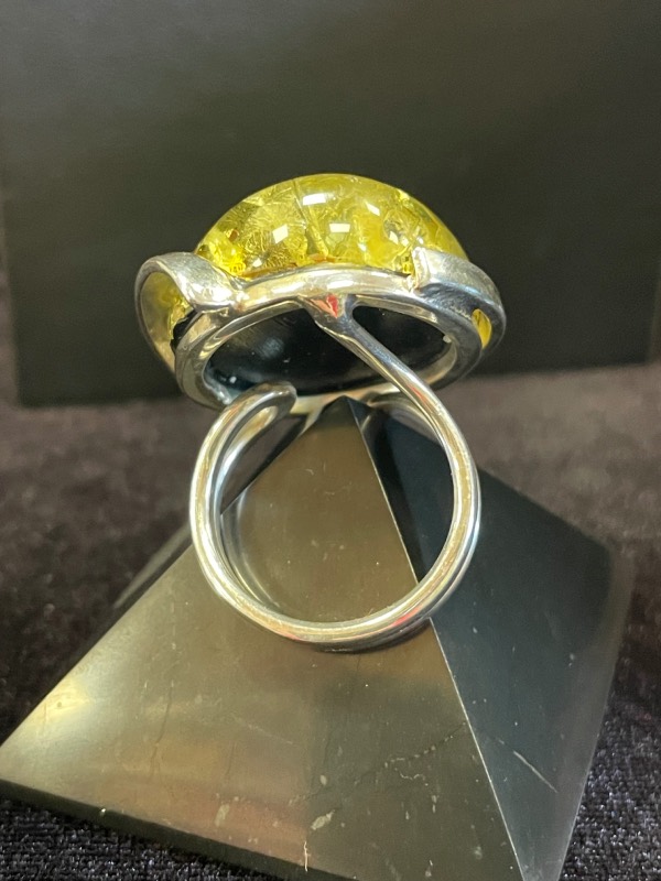 Bague ronde en argent avec une pierre minérale naturelle d’ambre vert de grosse taille. Montage atypique. Photo prise de derrière voire dessous sur fond noir avec la bague posée sur une pyramide de Shungite noire
