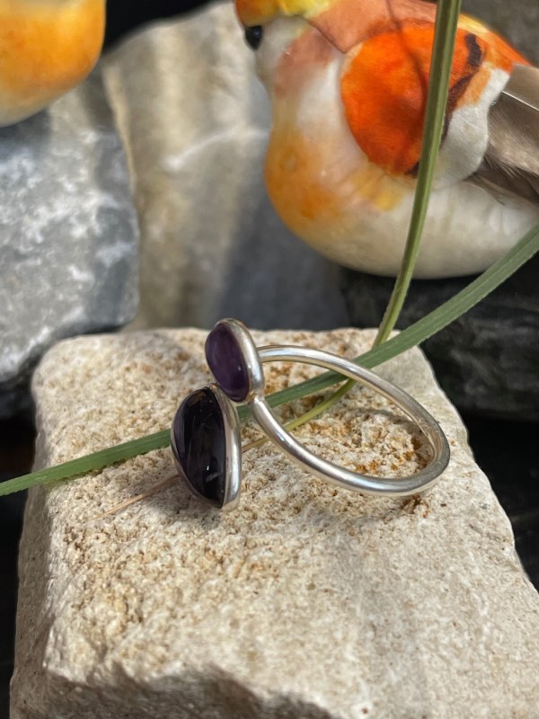 Bague en argent et 2 pierres d’améthyste violette, une plus grosse forme goutte et l’autre plus petite forme ovale. Bague posée sur pierre blanche décor nature . Photo prise de profil gauche