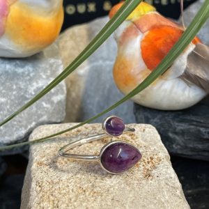 Bague en argent et 2 pierres d’améthyste violette, une plus grosse forme goutte et l’autre plus petite forme ovale. Bague posée sur pierre blanche décor nature . Photo prise de face
