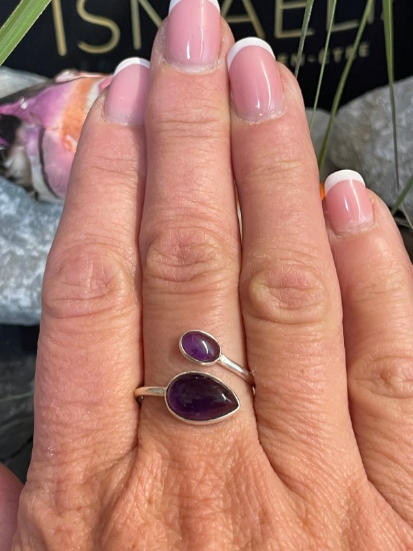 Bague en argent et 2 pierres d’améthyste violette, une plus grosse forme goutte et l’autre plus petite forme ovale. Bague portée sur une main. Photo prise de face