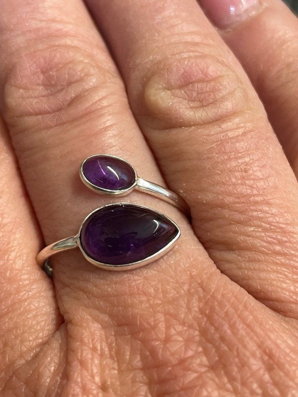 Bague en argent et 2 pierres d’améthyste violette, une plus grosse forme goutte et l’autre plus petite forme ovale. Bague portée sur une main. Photo prise de face Zoom sur la bague