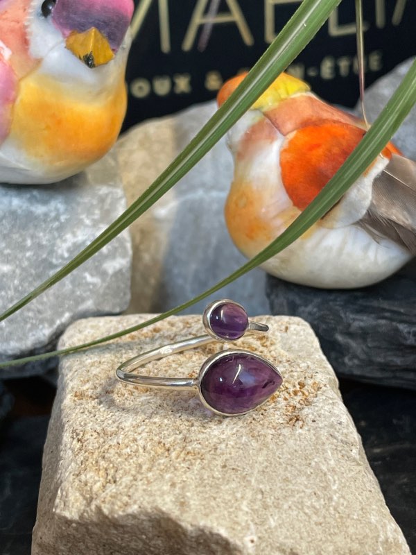 Bague en argent et 2 pierres d’améthyste violette, une plus grosse forme goutte et l’autre plus petite forme ovale. Bague posée sur pierre blanche décor nature . Photo prise de face
