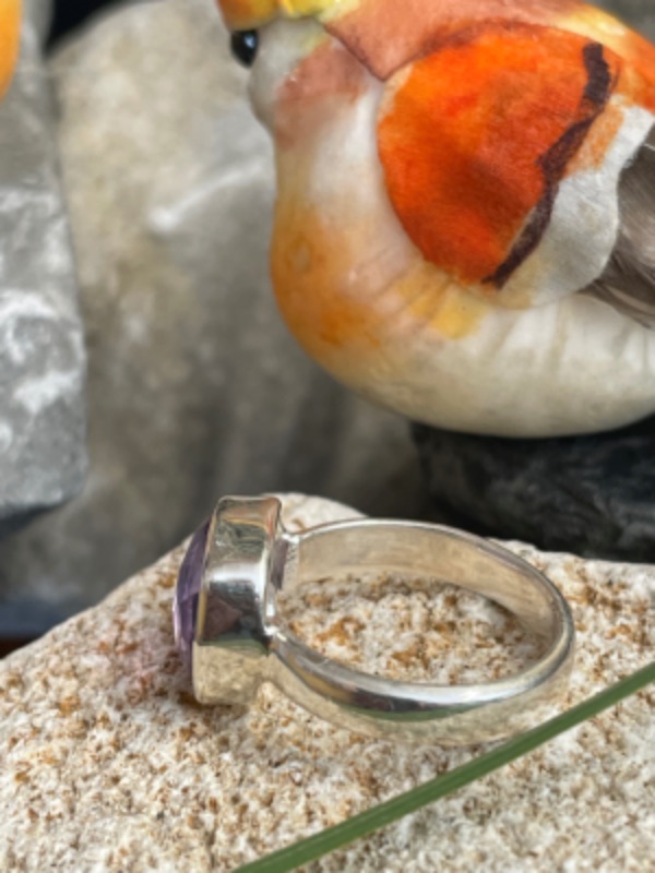 Bague en argent et pierre naturelle améthyste facettée de couleur violet clair transparent de forme carré. La bague est posée sur une pierre blanche avec un fond décor nature. Photo prise de profil gauche