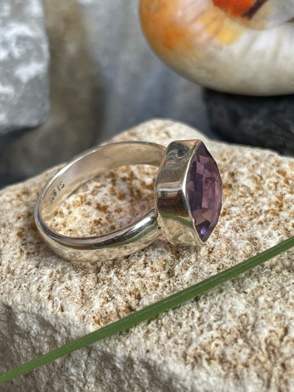 Bague en argent et pierre naturelle améthyste facettée de couleur violet clair transparent de forme carré. La bague est posée sur une pierre blanche avec un fond décor nature. Photo prise de profil droit
