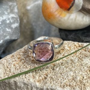 Bague en argent et pierre naturelle améthyste facettée de couleur violet clair transparent de forme carré. La bague est posée sur une pierre blanche avec un fond décor nature. Photo prise de face