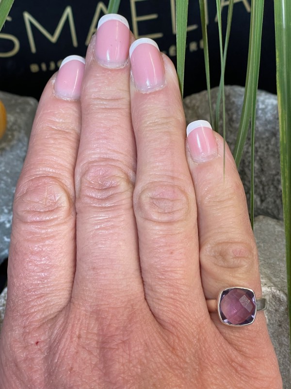 Bague en argent et pierre naturelle améthyste facettée de couleur violet clair transparent de forme carré. La bague est portée sur une main au petit doigt avec un fond décor nature. Photo prise de face