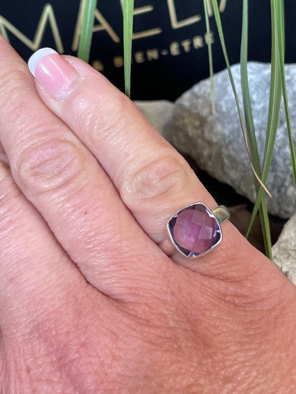 Bague en argent et pierre naturelle améthyste facettée de couleur violet clair transparent de forme carré. La bague est portée sur une main sur fond décor nature. Photo prise de face zoom sur le petit doigt