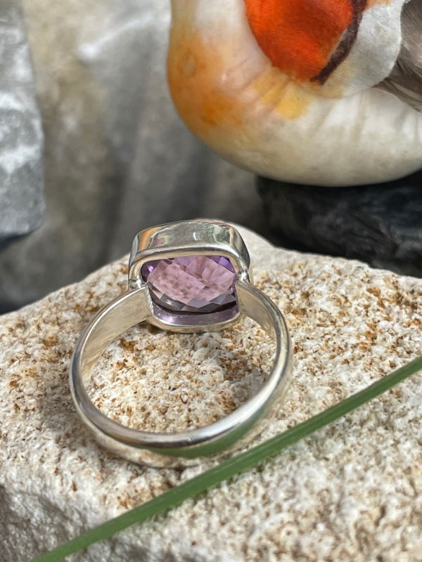 Bague en argent et pierre naturelle améthyste facettée de couleur violet clair transparent de forme carré. La bague est posée sur une pierre blanche avec un fond décor nature. Photo prise de l’arrière montrant le dessous de la pierre