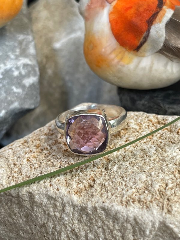 Bague en argent et pierre naturelle améthyste facettée de couleur violet clair transparent de forme carré. La bague est posée sur une pierre blanche avec un fond décor nature. Photo prise de face