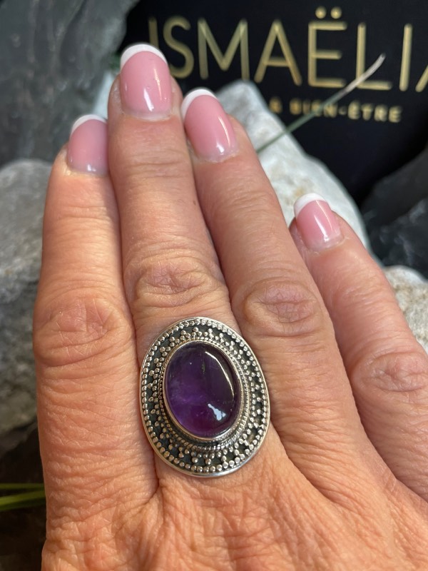 Bague en argent style ethnique et pierre améthyste violette de forme ovale. La bague est portee sur une main. Fond : pierres noires et blanches avec un peu de verdure. Main prise de face