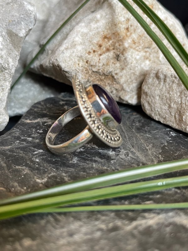 Bague en argent style ethnique et pierre améthyste violette de forme ovale. La bague est posée sur des pierres noires et blanches avec un peu de verdure. Bague prise de profil droit