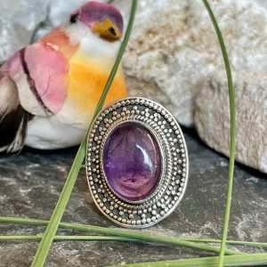 Bague en argent style ethnique et pierre améthyste violette de forme ovale. La bague est posée sur des pierres noires et blanches avec un peu de verdure. Bague prise de face