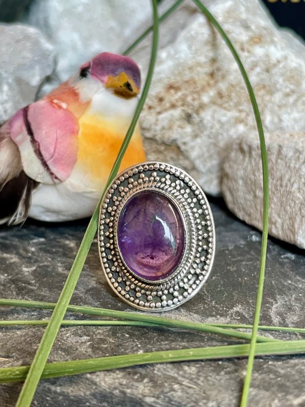 Bague en argent style ethnique et pierre améthyste violette de forme ovale. La bague est posée sur des pierres noires et blanches avec un peu de verdure. Bague prise de face