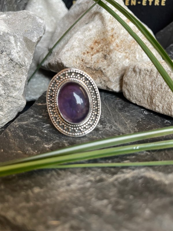 Bague en argent style ethnique et pierre améthyste violette de forme ovale. La bague est posée sur des pierres noires et blanches avec un peu de verdure. Bague prise de face