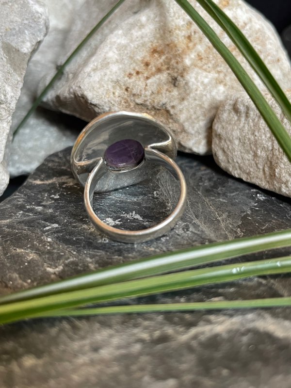 Bague en argent style ethnique et pierre améthyste violette de forme ovale. La bague est posée sur des pierres noires et blanches avec un peu de verdure. Bague prise de l’arrière pour voir le dessous de la pierre