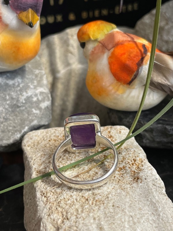 Bague argent et pierre améthyste rectangle posée sur une pierre blanche décor nature. Photo prise de l’arrière pour voir le montage sous la pierre