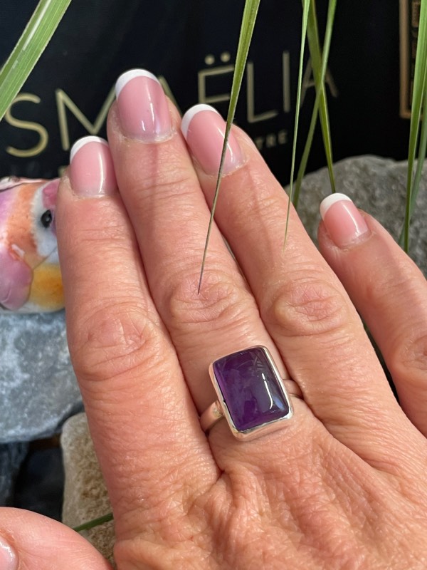 Bague argent et pierre améthyste rectangle portée sur une main, fond décor nature. Photo prise de face
