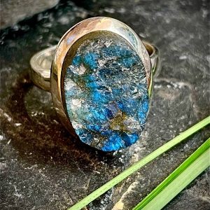 Bague argent 925 et pierre naturelle labradorite brute grise a reflets bleus de forme ovale. Bague posée sur une pierre noire sur fond nature. Photo prise de face