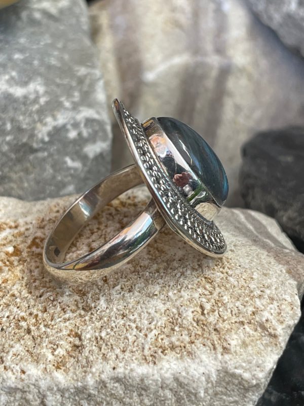 Bague en argent et pierre naturelle labradorite de forme ovale décorée style ethnique autour. La bague est posée sur une pierre blanche sur fond décor naturel. Photo prise de profil droit