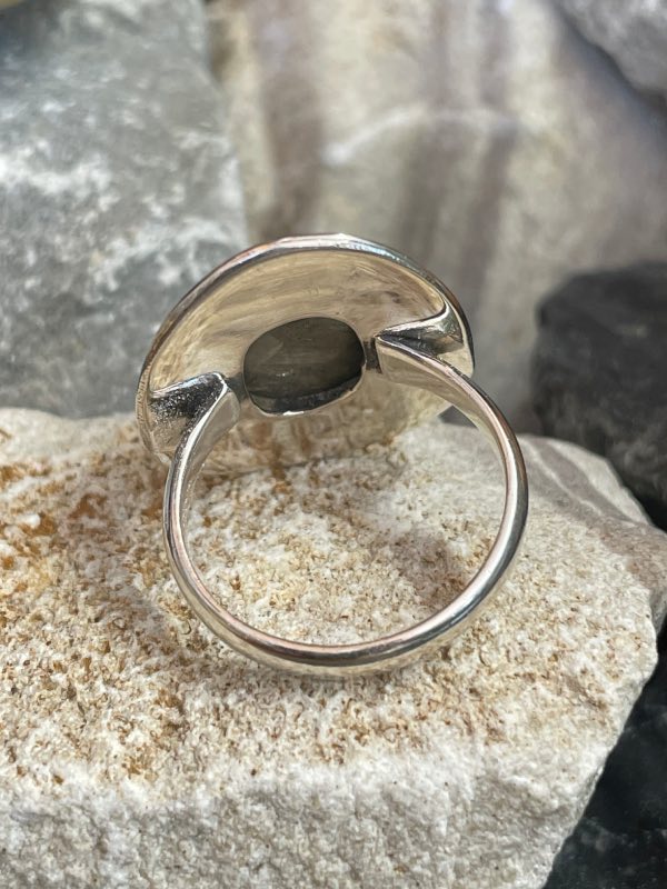 Bague en argent et pierre naturelle labradorite de forme ovale décorée style ethnique autour. La bague est posée sur une pierre blanche sur fond décor naturel. Photo prise de l’arrière pour montrer le dessous de la pierre