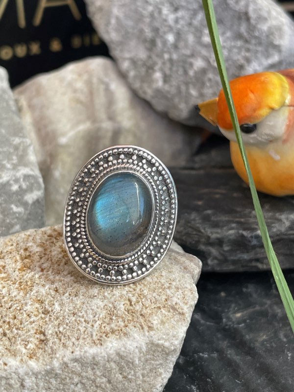 Bague en argent et pierre naturelle labradorite de forme ovale décorée style ethnique autour. La bague est posée sur une pierre blanche sur fond décor naturel. Photo prise de face