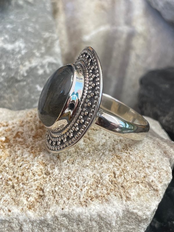 Bague en argent et pierre naturelle labradorite de forme ovale décorée style ethnique autour. La bague est posée sur une pierre blanche sur fond décor naturel. Photo prise de profil gauche