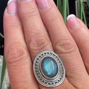 Bague en argent et pierre naturelle labradorite de forme ovale décorée style ethnique autour. La bague est portée sur une main sur fond décor naturel. Photo prise de face zoomé