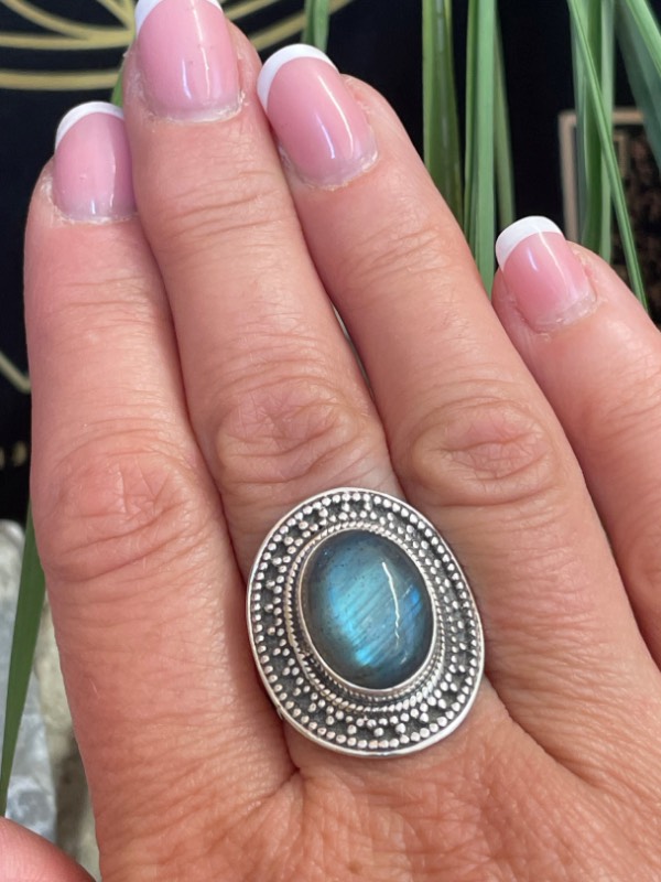 Bague en argent et pierre naturelle labradorite de forme ovale décorée style ethnique autour. La bague est portée sur une main sur fond décor naturel. Photo prise de face zoomé