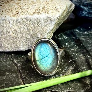 Bague en argent 925 et pierre naturelle labradorite de forme ovale taille moyenne posée sur une pierre noire sur fond nature. Photo prise de face