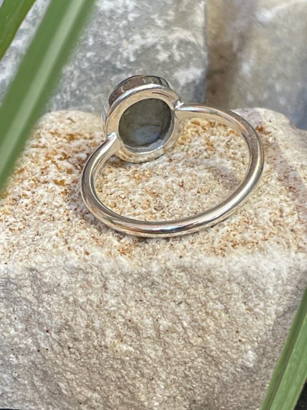 Bague en argent 925 et labradorite de couleur bleu/vert clair de forme ovale. Elle est posée sur une pierre blanche avec un fond naturel. La photo est prise de l’arrière pour montrer sous la pierre