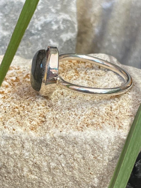 Bague en argent 925 et labradorite de couleur bleu/vert clair de forme ovale. Elle est posée sur une pierre blanche avec un fond naturel. La photo est prise de profil gauche