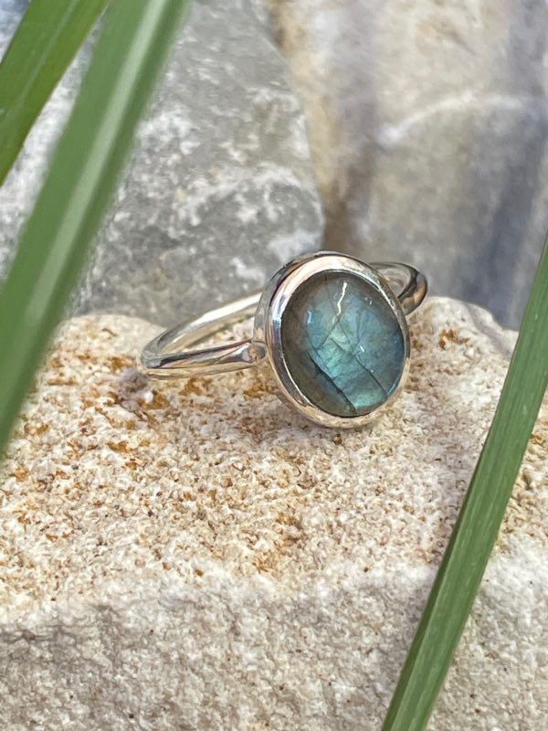 Bague en argent 925 et labradorite de couleur bleu/vert clair de forme ovale. Elle est posée sur une pierre blanche avec un fond naturel. La photo est prise de face