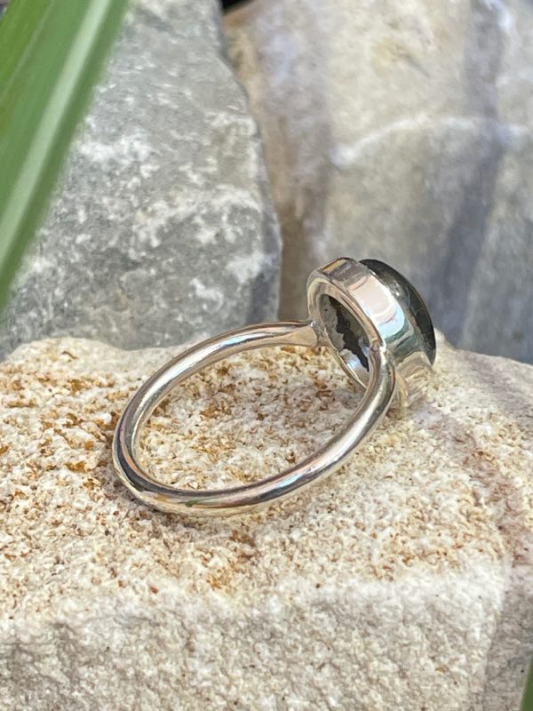 Bague en argent 925 et labradorite de couleur bleu/vert clair de forme ovale. Elle est posée sur une pierre blanche avec un fond naturel. La photo est prise de profil droit